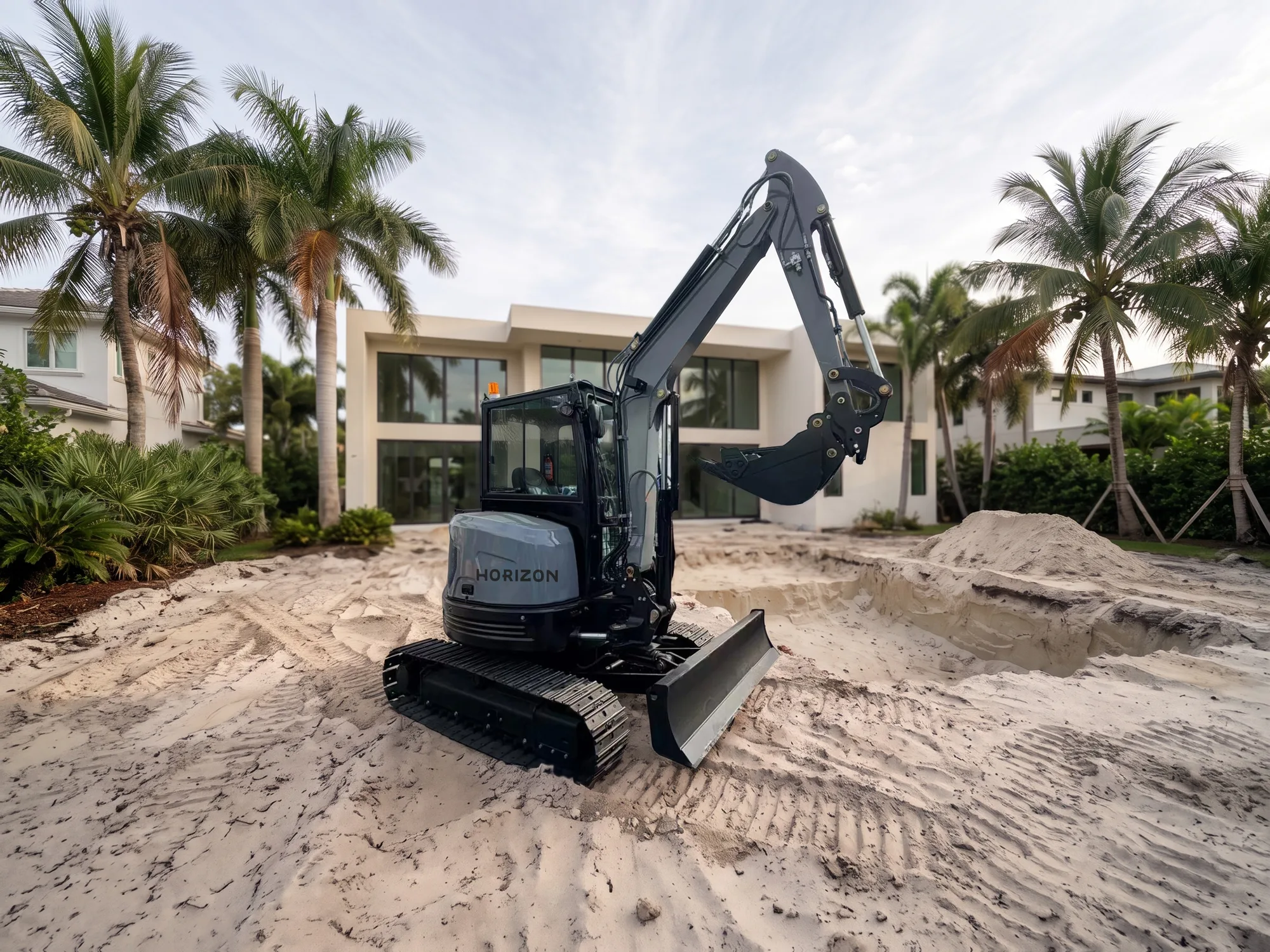 Miniexcavadora gris con cabina negra cerrada, hoja niveladora y cuchara, HORIZON en el contrapeso, excavando arena en una obra residencial en el sur de Florida